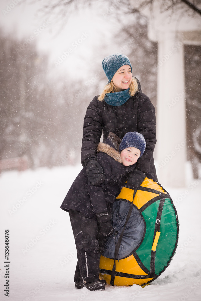Fototapeta premium Young smiling woman and cute kid boy with inflatable sled walking in a winter park. Happy family enjoying snow walk. Mother and son. Lifestyle concept