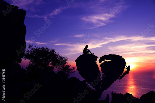 Women sitting and Men climbing on broken heart-shaped stone on a mountain with purple sky sunset background.Silhouette Valentine background concept.