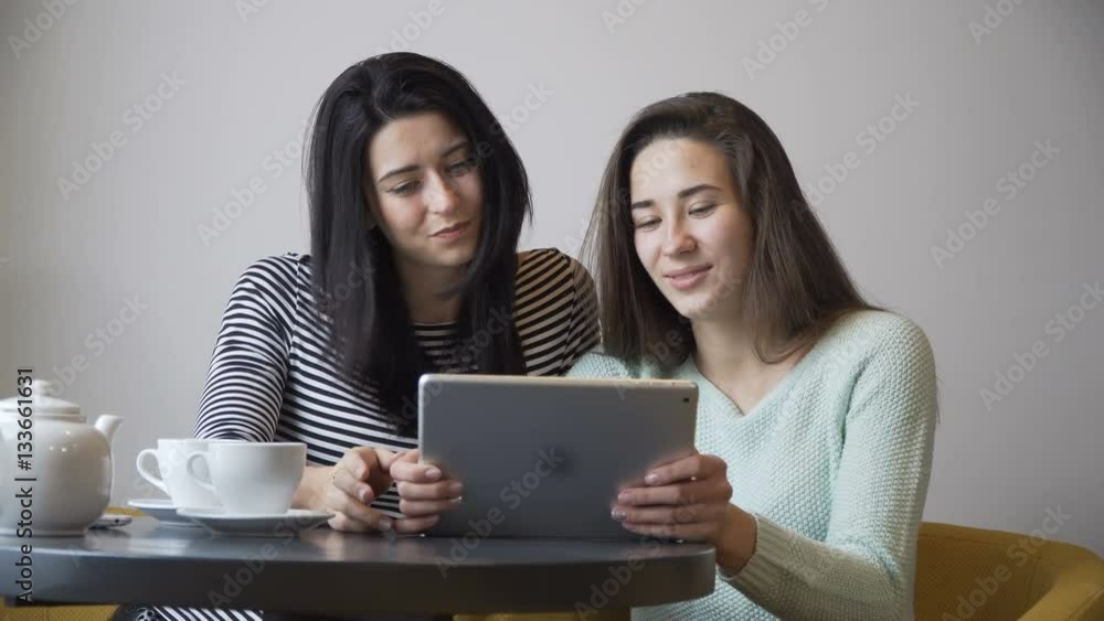 Two beautiful girls sitting in cafe, looking a tablet PC and laugh. Camera spies. Two beautiful girls sitting in cafe, looking a tablet PC and laugh. Camera spies.