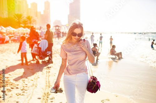 Photography Thin blonde lady holds her shoes walking along sand beach in Dub