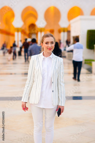 Pretty blonde woman in white suit and beige shoes stands on sunn