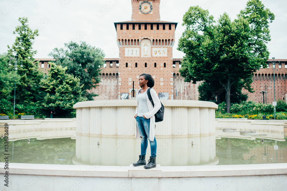 Young beautiful afro woman posing in front of Castello Sforzesco in the city center of Milan, overlooking smiling - traveler, student, happiness concept