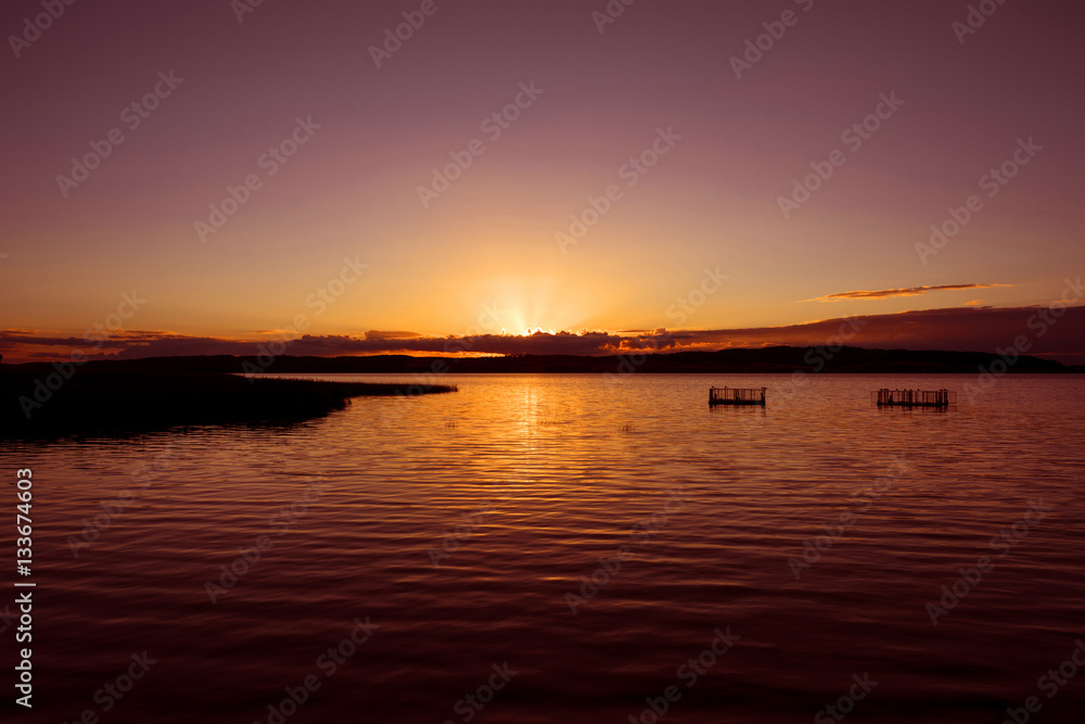 Germany, Kummerow: Atmospheric colorful sunset with lake, sunbeams, weirs, reed, horizon and sky.