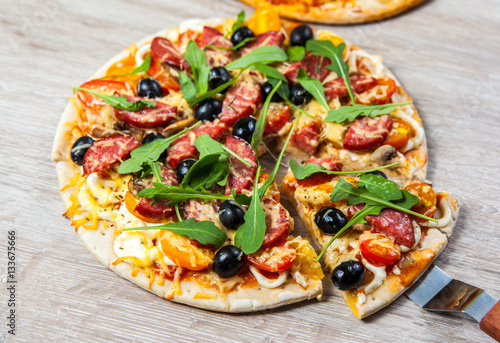 Cook takes a shovel one piece of pizza with cheese olives and salami, mushrooms and tomatoes, arugula. On a wooden stand