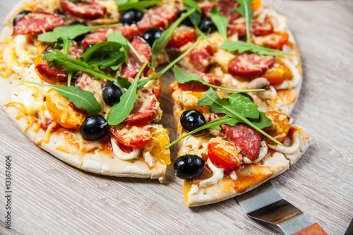 Cook takes a shovel one piece of pizza with cheese olives and salami, mushrooms and tomatoes, arugula. On a wooden stand