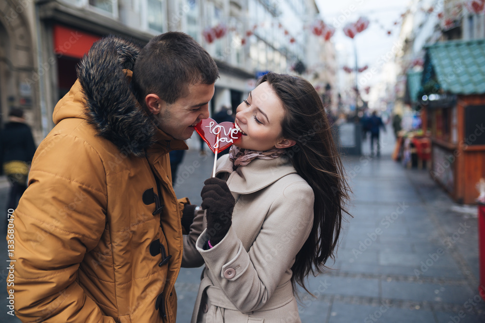 Fototapeta premium Beautiful young couple in love enjoying together at city street and eating together a lollipop in heart shape.