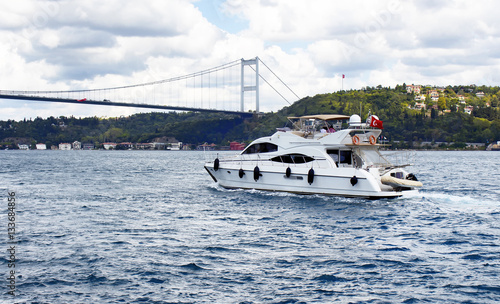 White luxury yacht / boat crosses Bosphorus between European and Asian sides in Istanbul. It is cloudy autumn day. FSM (Fatih Sultan Mehmet) bridge is in the background.