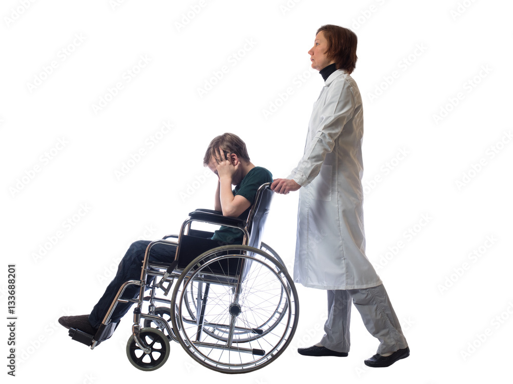 Young distressed boy sitting in a wheelchair holding his head and behind a wheelchair stands a woman in the medical gown on a white background Young distressed boy sitting in a wheelchair holding his head and behind a wheelchair stands a woman in the medical gown on a white background