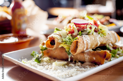 Gourmet Rolled Chicken and Beef Taquitos with Mozzarella Cheese, Lettuce, Sliced Red Beet and Tomato Garnish, Green and Red Tomatillo Sauce and other mexican food blurred in the background photo