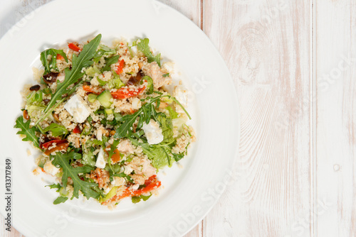 Couscous salad on a white plate placed on white wood. The salad ingredients consist of rocket, feta cheese, pine nuts and roasted peppers. 