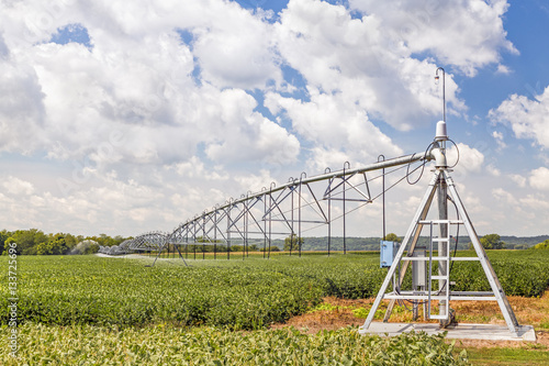 A soybean field in the American Midwest is watered by a center pivot irrigation system under a cloudy blue sky.