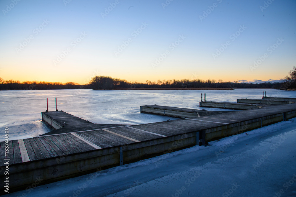Fototapeta premium wide angle shot of wooden boat docks on icy blue frozen lake with warm orange and yellow sunset on the horizon