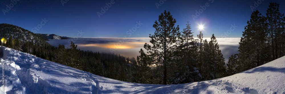 Naklejka premium Cold Winter Snowy landscape at night with cloud inversion covering city lights that glow underneath the cloud cover. Lit with moonlight and the sky has stars.