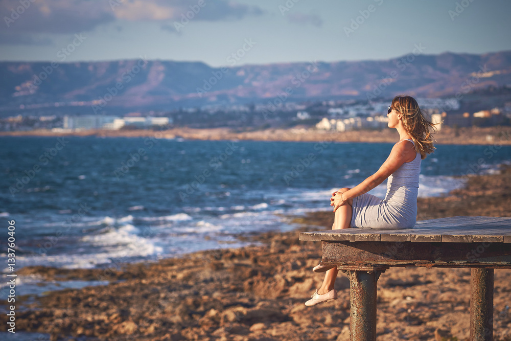 Young woman with long hair sitting at the edge of wooden pier and watching sunset at the rocky coast of Paphos, Cyprus