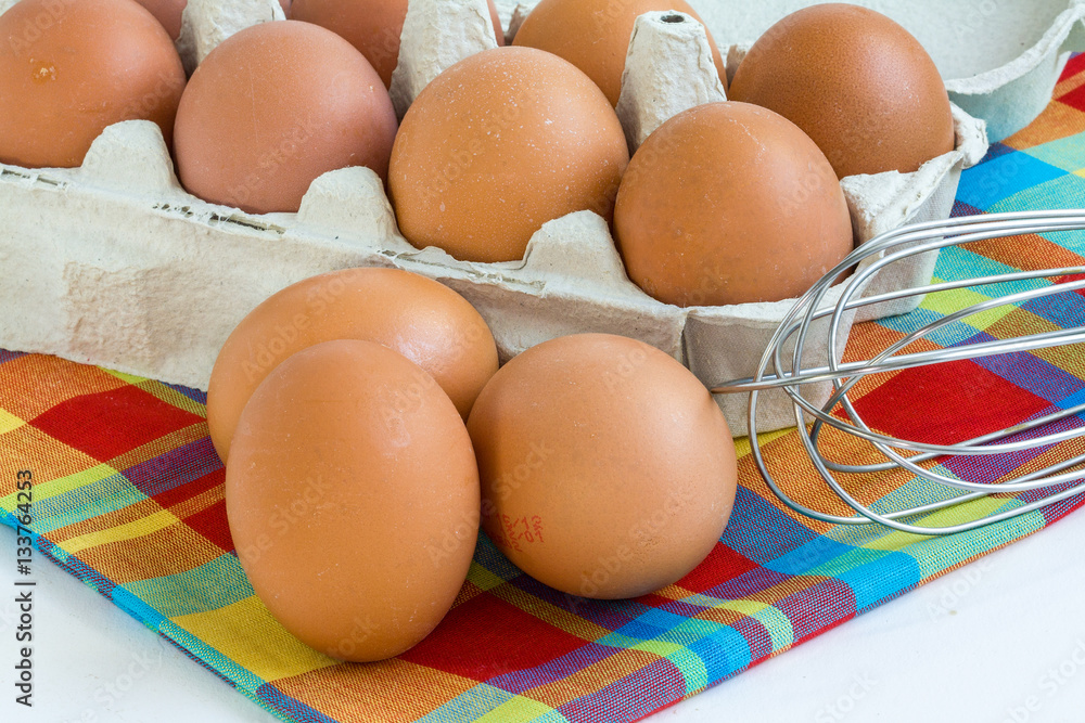 Oeufs dans leur boîte et batteur manuel sur fond de tissu madras Oeufs dans leur boîte et batteur manuel sur fond de tissu madras