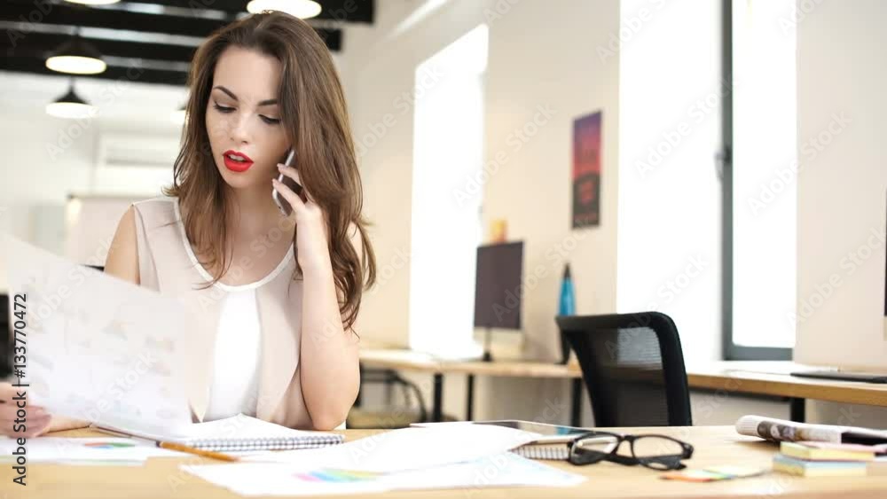 Young business woman talking on mobile phone and making notes in notebook while sitting at the table in office Young business woman talking on mobile phone and making notes in notebook while sitting at the table in office