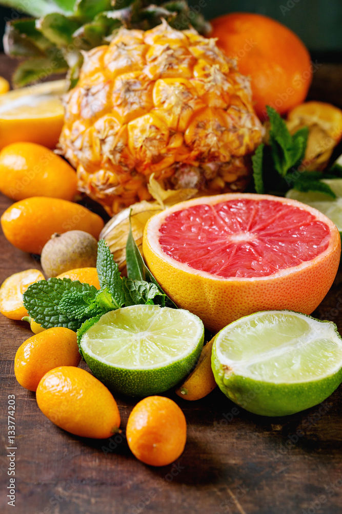 Variety of whole and sliced citrus fruits pineapple, grapefruit, lemon, lime, kumquat, clementine and physalis with mint over dark wooden background. Top view with space. Healthy eating, dieting