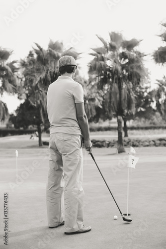 Back view of active man holding playing vintage wooden golf club on palm tree grass field outdoors background.