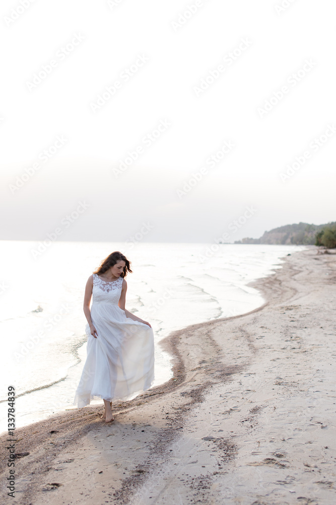 Beautiful young brunette woman in white long dress at the sea shore