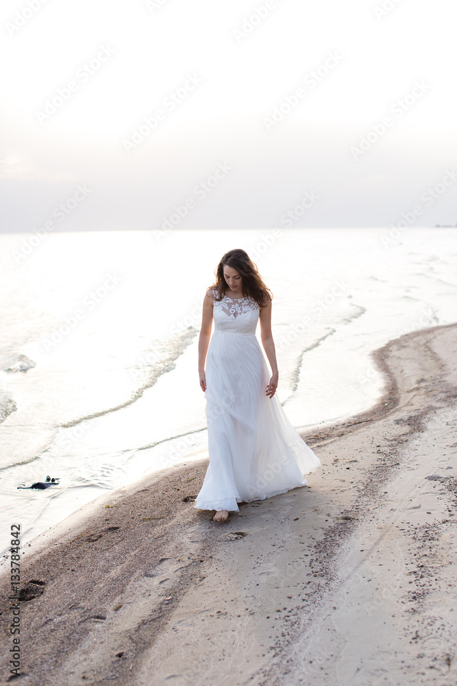 Beautiful young brunette woman in white long dress at the sea shore