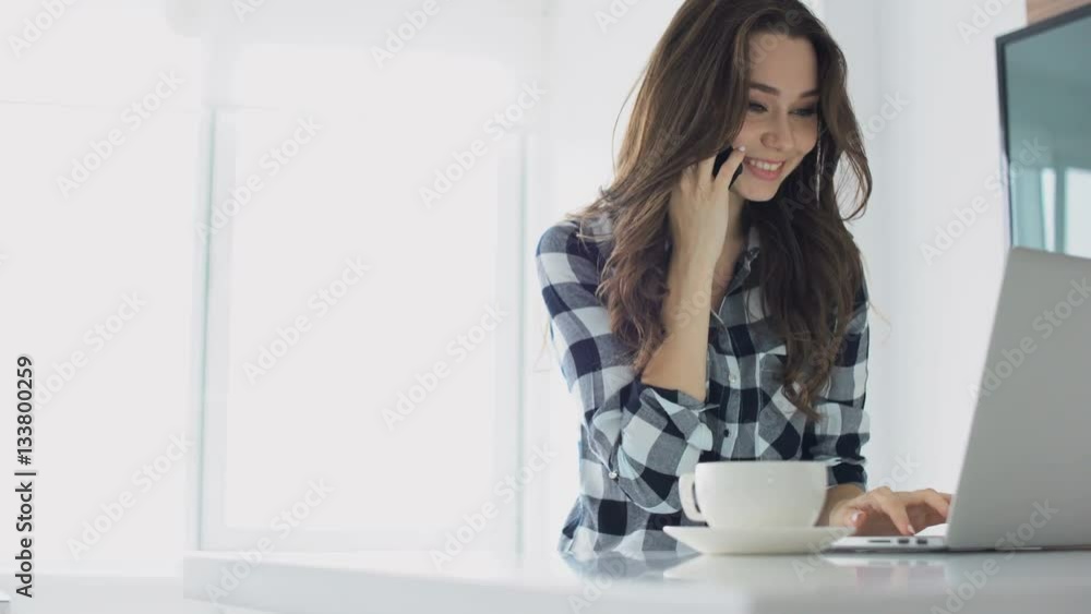 Smiling happy woman talking on mobile phone while using laptop at home Smiling happy woman talking on mobile phone while using laptop at home