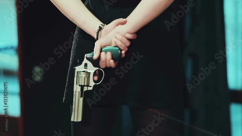 Young girl in a black dress and red shoes, holding a gun behind.