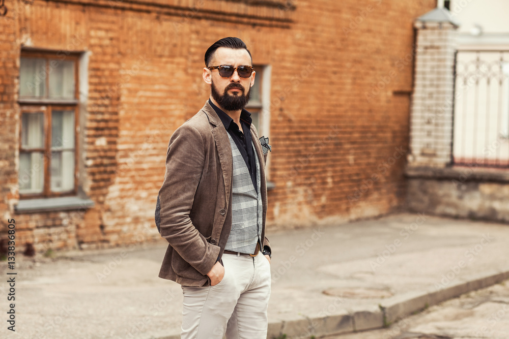 Young confident hipster man with beard in glasses posing on the street in  old town. Cute