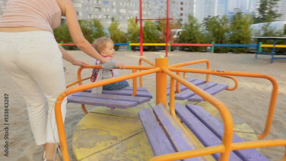 Beautiful young mother plays with her baby son on carousel at playground Beautiful young mother plays with her baby son on carousel at playground