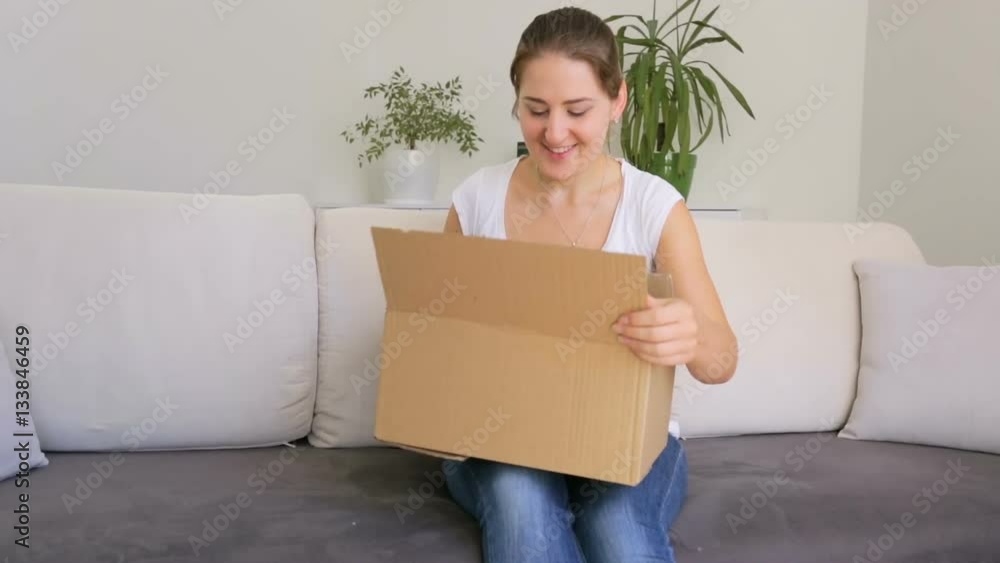 Happy young woman sitting on sofa at living room and taking dress out of box she bought at online store Happy young woman sitting on sofa at living room and taking dress out of box she bought at online store