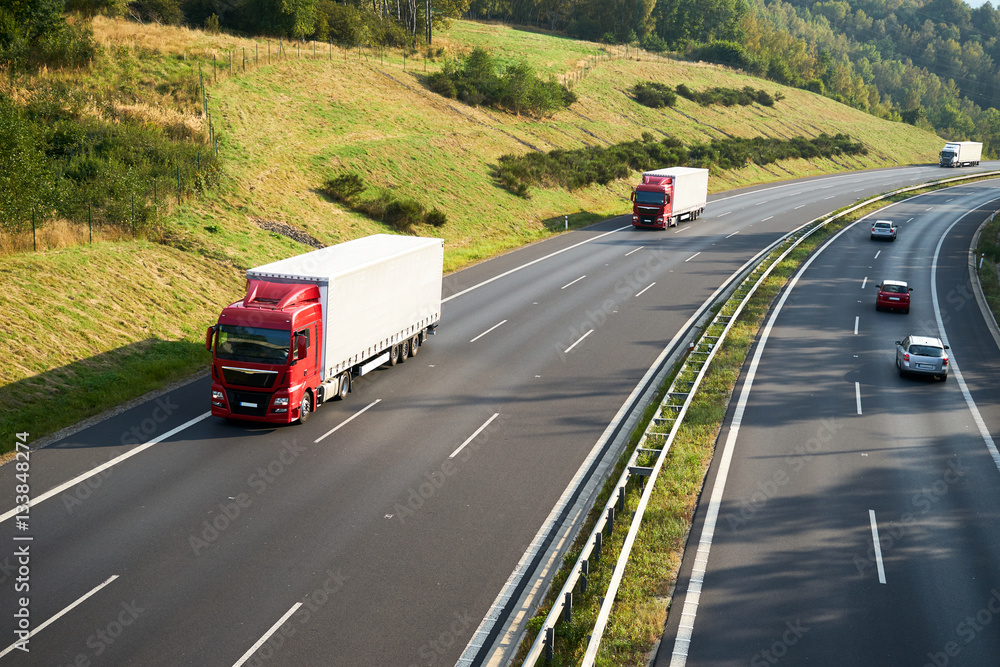 Three trucks and three cars driving on the highway beneath a hillside meadow and forest. View from above.