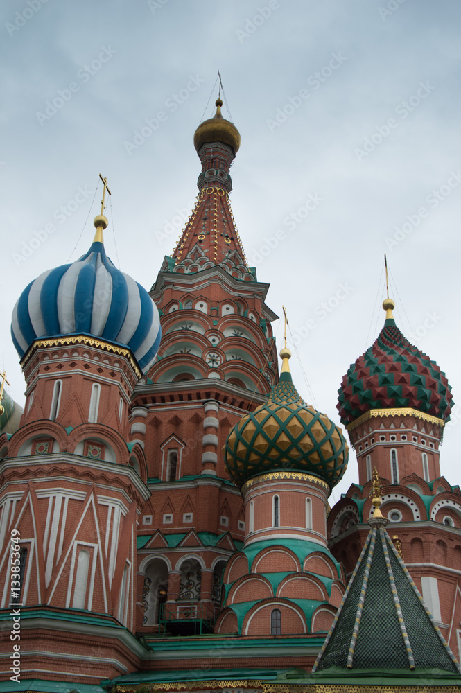 St. Basil's Cathedral exterior showing decorative red brick facade and colorful onion domes topped with Orthodox crosses against a blue sky with thin clouds. 