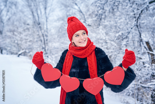 Love and valentines day concept. Smiling woman holding garland of four red paper hearts shape - blank copy space for letters or text, looking away over winter landscape