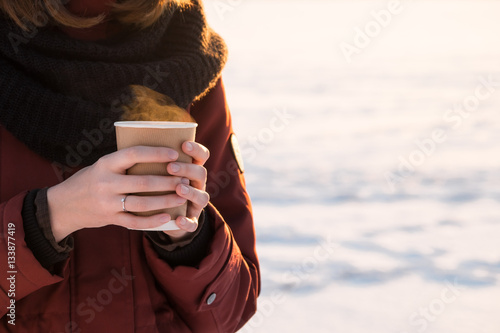 Person with cup of hot coffee outdoors at sunset. Girl in red parka coat and scarf holding hot steaming drink outdoors on a sunny winter day photo