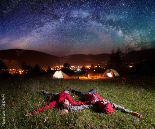 Twosome admiring the bright stars and lying on the grass in the background night tent camping near campfire, mighty mountains, luminous town