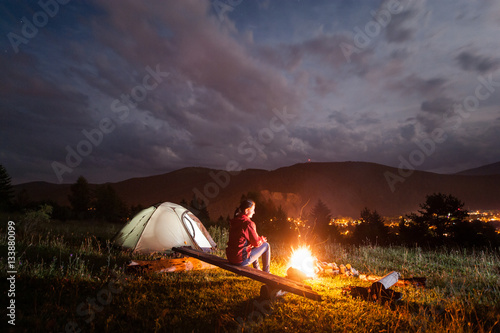 Woman sitting on boards enjoying a bonfire near tent under cloudy sky in the background mountains and luminous town in the evening photo