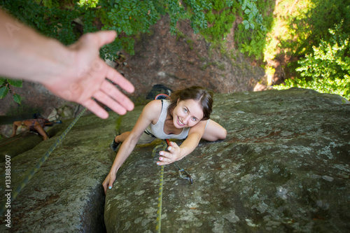 Young happy female climber is climbing on high boulder and try to catch helping hand. Top view
