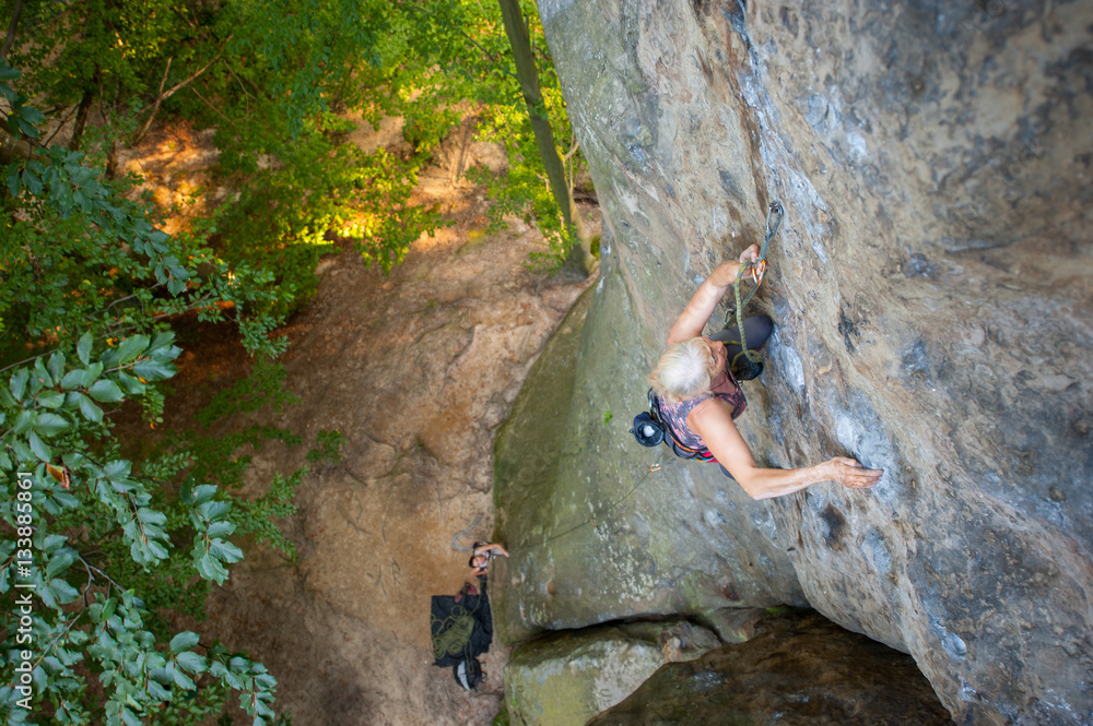 Obraz premium Older woman rock climber is climbing with carbines and rope on a rocky wall of big rock. Belayer standing on the ground insuring the climber. View from the top. Extreme sports concept