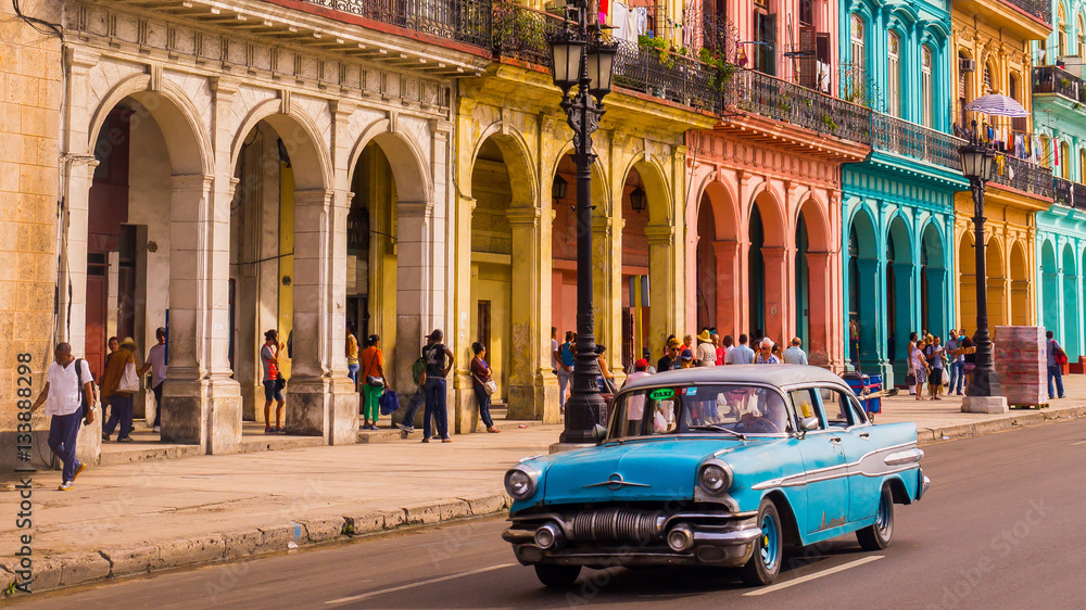 Naklejka premium Niebieska taksówka weterana przejeżdża przez Habana Vieja przed kolorową fasadą