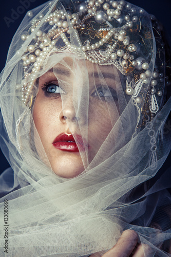 Φωτογραφία fashion beauty portrait of young beautiful young woman with makeup and freckles on her face and pearl headpiece on her head and white tulle in front of her face on dark blue background
