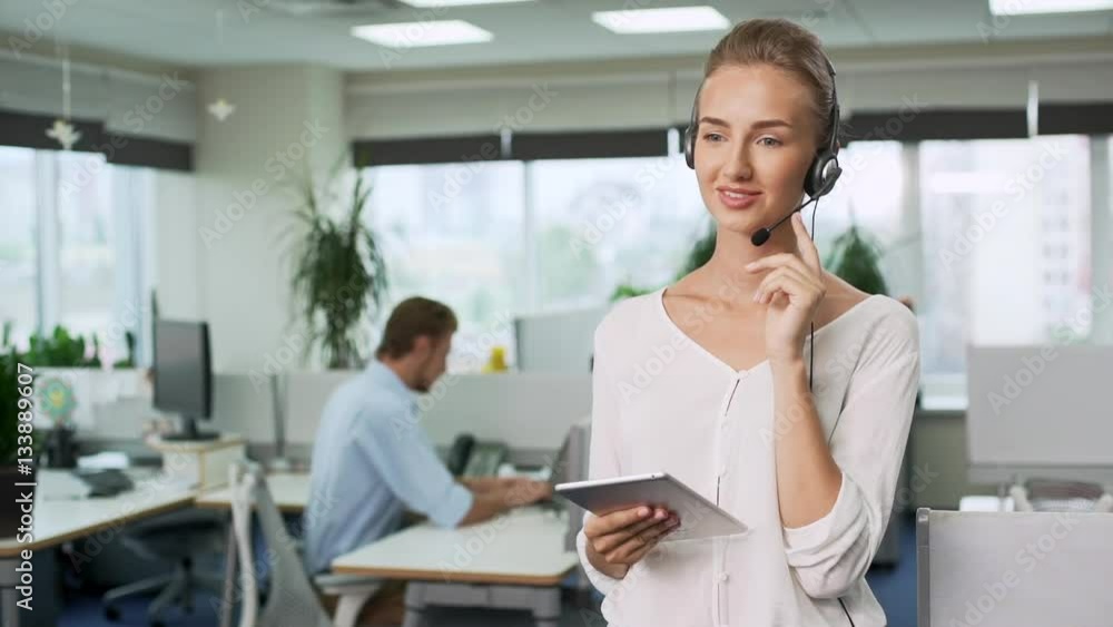 Young beautiful businesswoman in headset holding tablet showing thumb up coworker working on background Slow motion Young beautiful businesswoman in headset holding tablet showing thumb up coworker working on background Slow motion