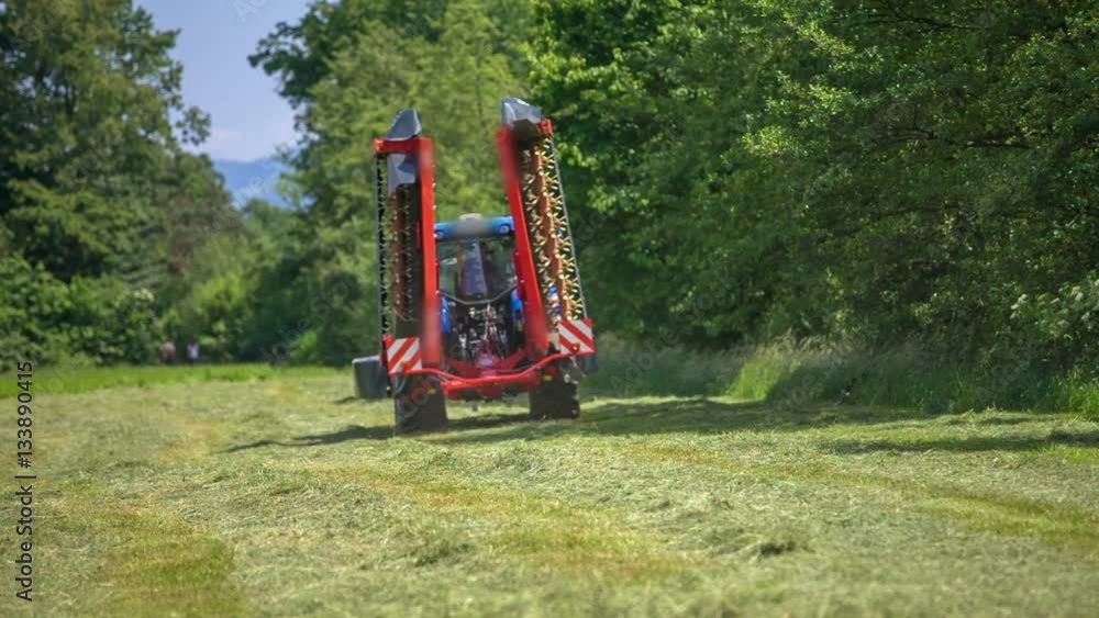 A big tractor is leaving a freshly mowed lawn. There is a machinery connected to it. It is a nice summer day.
