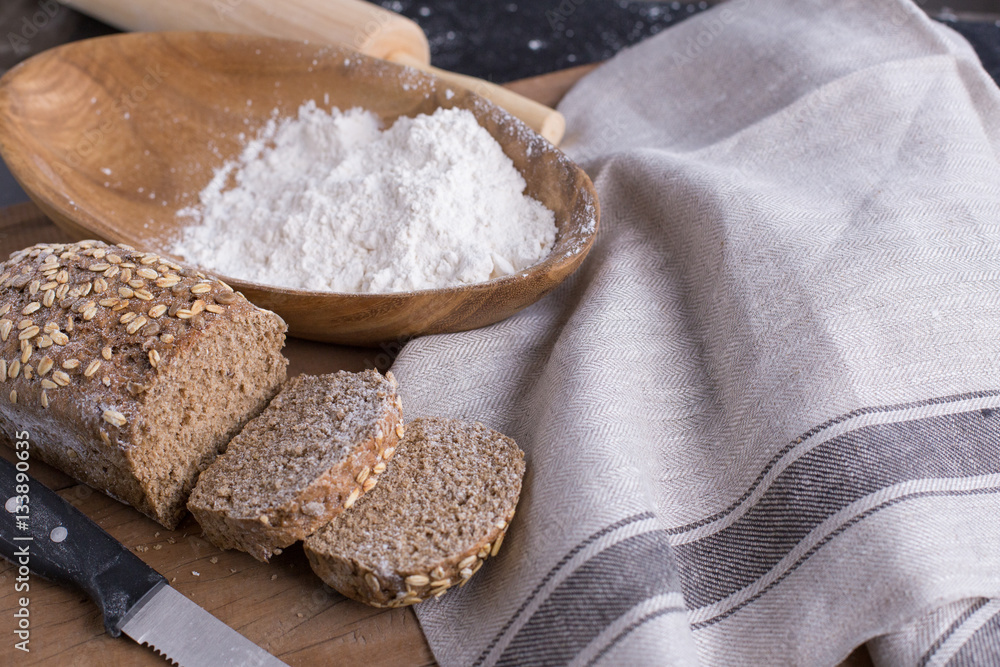 Cut whole grain wheat bread in pieces set on nice fabric brown background decorated with flour bowl and a bakery wooden roller.