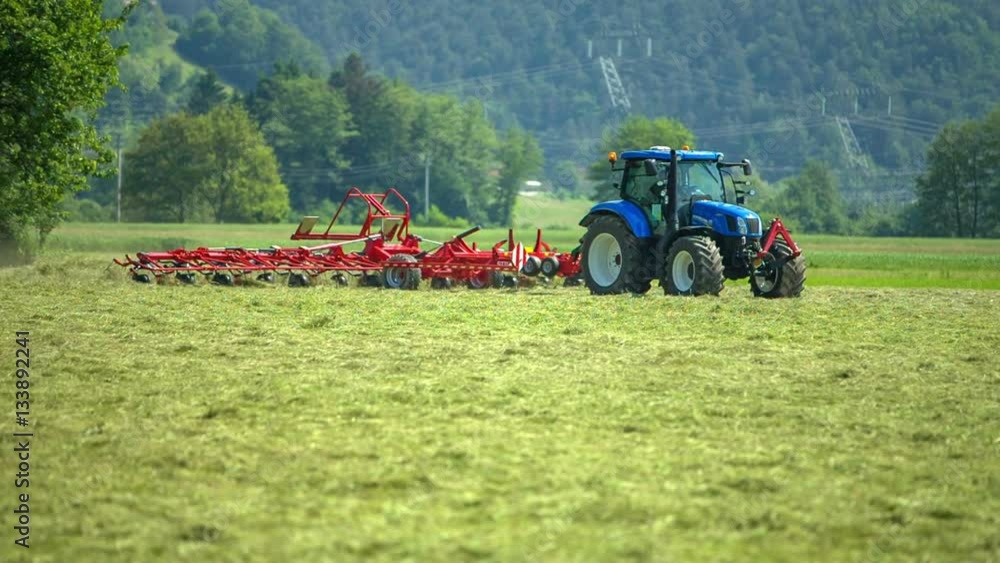 A blue tractor is preparing hay. With the help of the agricultural machinery it is turning hay around.
A blue tractor is preparing hay. With the help of the agricultural machinery it is turning hay around.