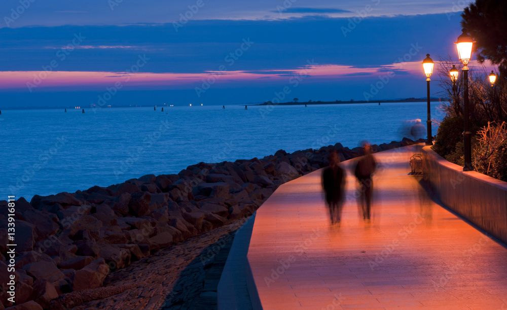 Obraz premium People walk on the seafront in little town of Grado at sunset. Italy.