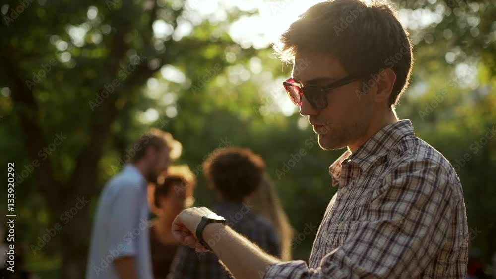 Caucasian male in plaid shirt and sunglasses standing in park using tablet checking time on wrist watch in slowmotion Caucasian male in plaid shirt and sunglasses standing in park using tablet checking time on wrist watch in slowmotion