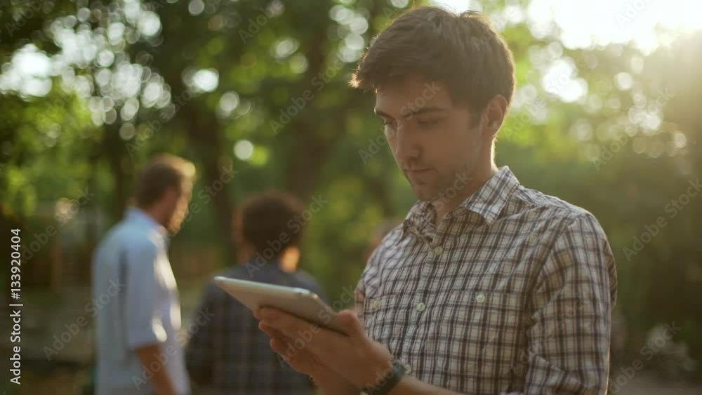 Young handsome Caucasian male in plaid shirt standing in park with tablet showing ok with thumb with people chatting on background in slowmotion Young handsome Caucasian male in plaid shirt standing in park with tablet showing ok with thumb with people chatting on background in slowmotion