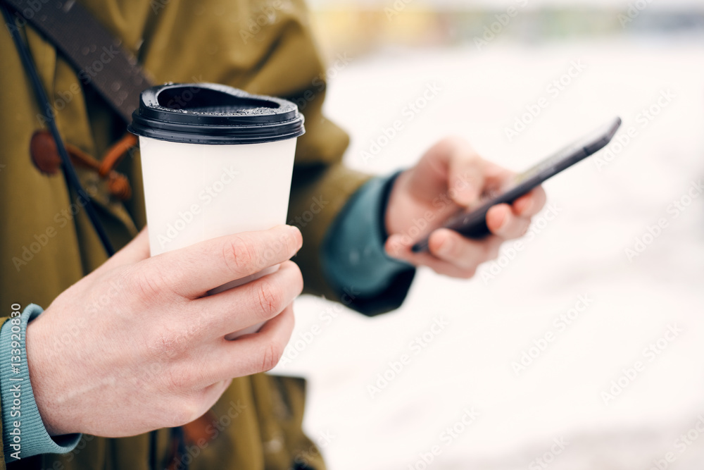 Man holding a cup of coffee in their hands and looking at phone on the street in winter Man holding a cup of coffee in their hands and looking at phone on the street in winter