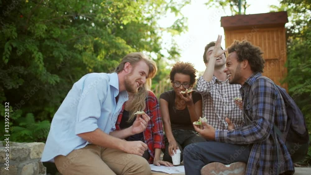 Five interracial friends actively speaking laughing smiling eating pizza in park in slowmotion