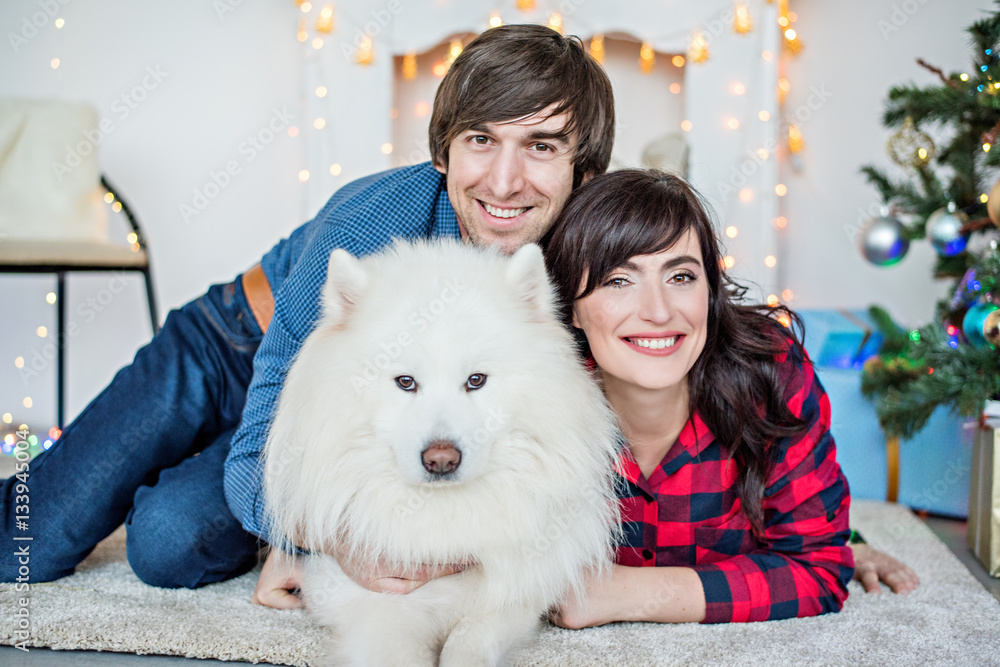 At Christmas, a large portrait of a beautiful couple in love by a white fireplace hugs a Samoyed husky. Against the background of festive yellow blurry lights and gifts at the Christmas tree. New Year At Christmas, a large portrait of a beautiful couple in love by a white fireplace hugs a Samoyed husky. Against the background of festive yellow blurry lights and gifts at the Christmas tree. New Year