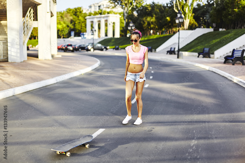 Beautiful hipster girl in a jeans shorts and pink shirt walking with skateboard near the green park, sexy style, adult, long legs, brunette, white shoes, sexy model, posing on camera